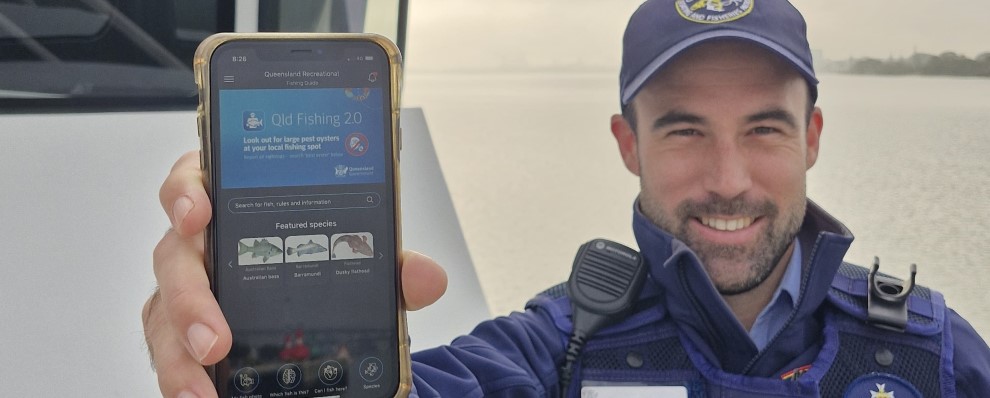 Queensland Boating and Fisheries Patrol officer in uniform, standing on a boat in water, holding a mobile phone to the camera, showing the Qld fishing 2.0 app.